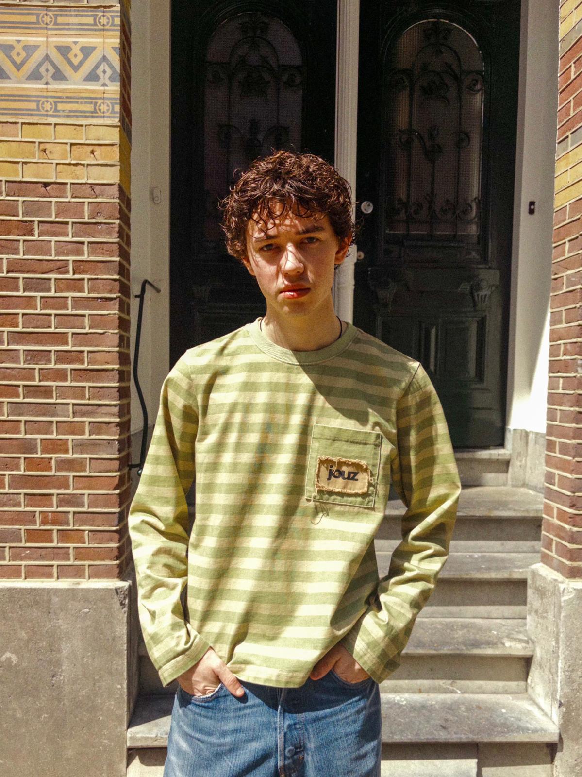 Person wearing a green striped shirt with a logo, standing in front of a brick building.