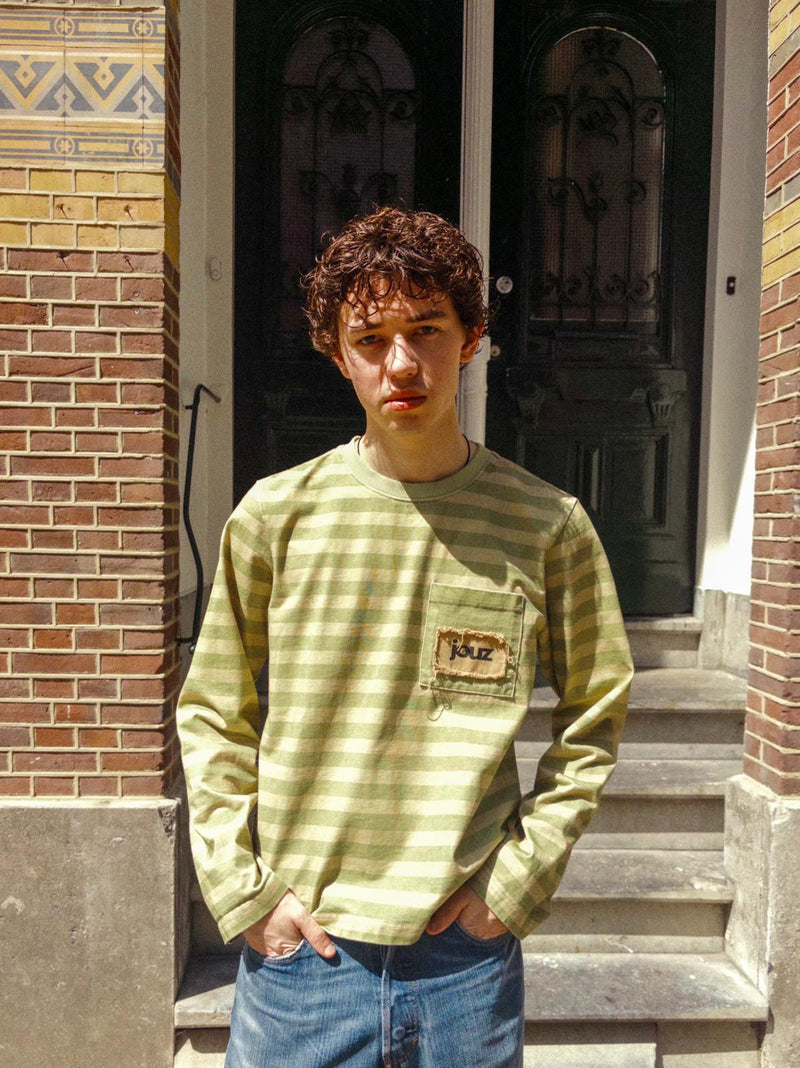 Person wearing a green striped shirt with a logo, standing in front of a brick building.