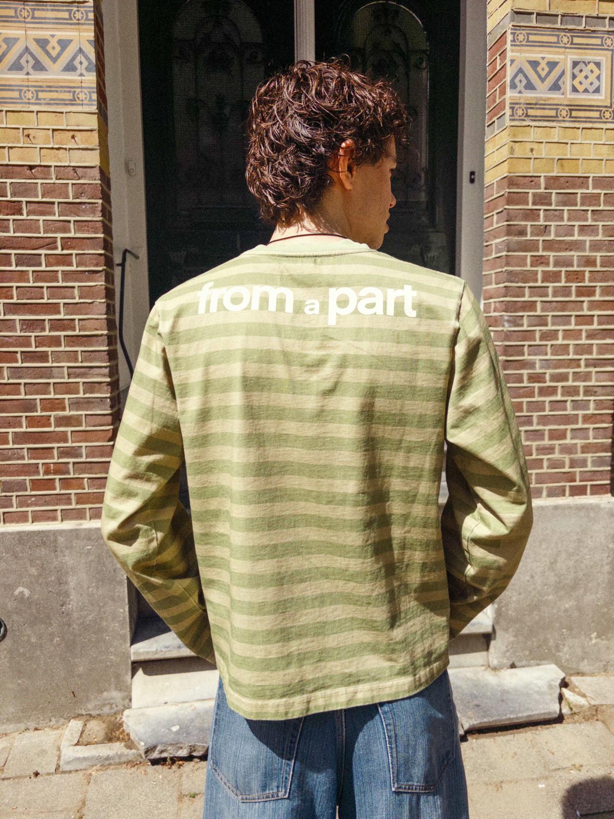 Person wearing a green shirt with 'from part' text on the back, standing in front of a brick building.